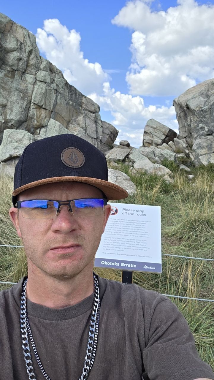 Derick in a black cap and glasses, in front of the fence and the 'Please stay off the rocks' sign; the rock's split rises behind and above him against blue sky and cloud.