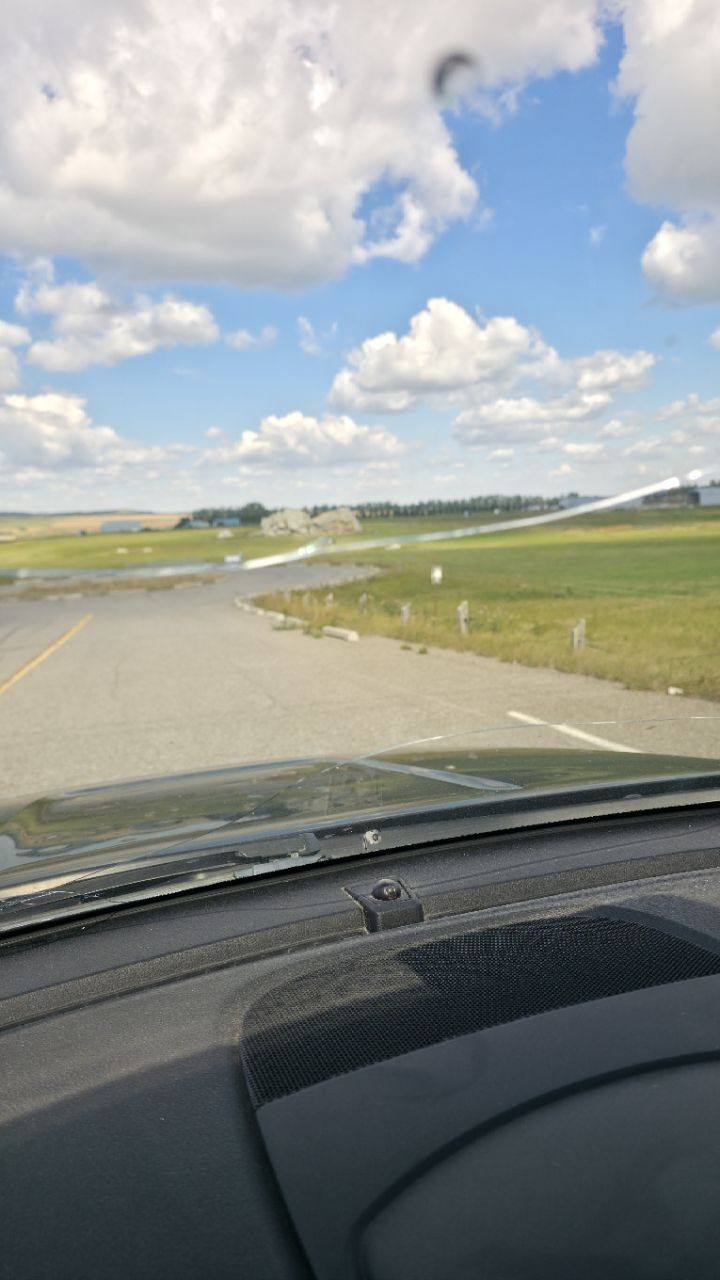 Through a windshield: a wide grey lot and an access road, then flat green fields with farm buildings, and in the middle distance a lone pale grey boulder the size of a house standing on open ground under a big cumulus sky.