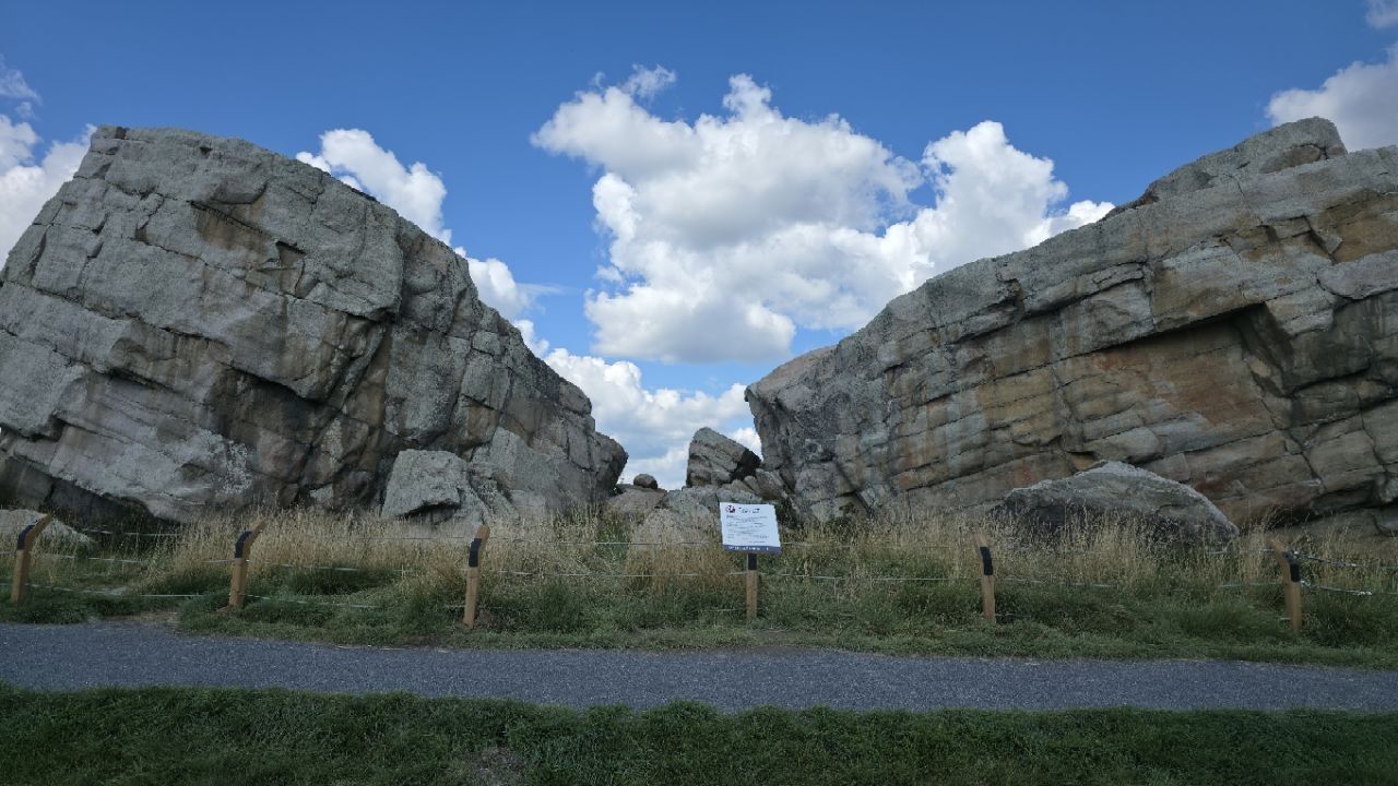 Two enormous angular quartzite blocks, each several storeys high, with a gap between them full of sky and smaller broken blocks; a low post-and-cable fence and a small interpretive sign in the foreground; a gravel path.
