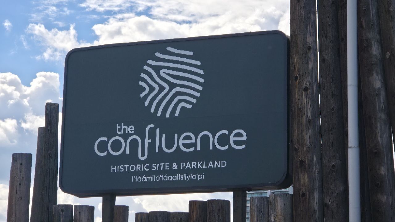 A dark green sign on wooden palisade posts against cloud: a fingerprint-like spiral logo, then 'the confluence', then 'HISTORIC SITE & PARKLAND', then the Blackfoot name I'táámito'táaattsiiyio'pi.