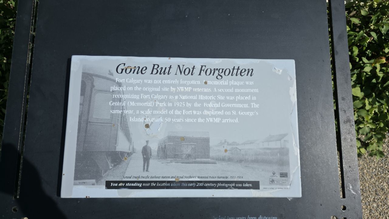 A weathered grey plaque titled 'Gone But Not Forgotten', with an early-20th-century photograph of a railway station and barracks, and text about memorial plaques placed on the site by NWMP veterans.