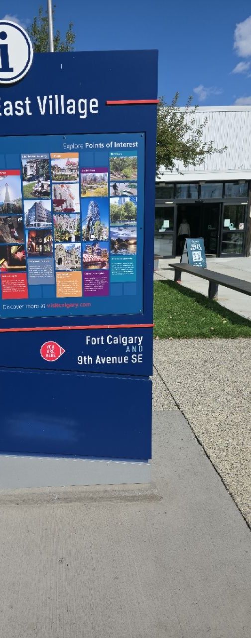 A blue City of Calgary East Village wayfinding pillar with a points-of-interest board; below it a red 'You are here' marker beside the words 'Fort Calgary AND 9th Avenue SE'.