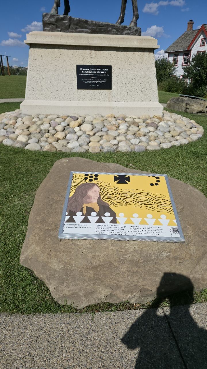 The stone base of the Colonel James Macleod equestrian statue, ringed with river rocks; in front of it a large flat boulder carrying a framed 2023 artwork of Crowfoot in yellow and black by Sikapinakii Low Horn; the photographer's shadow across the grass.
