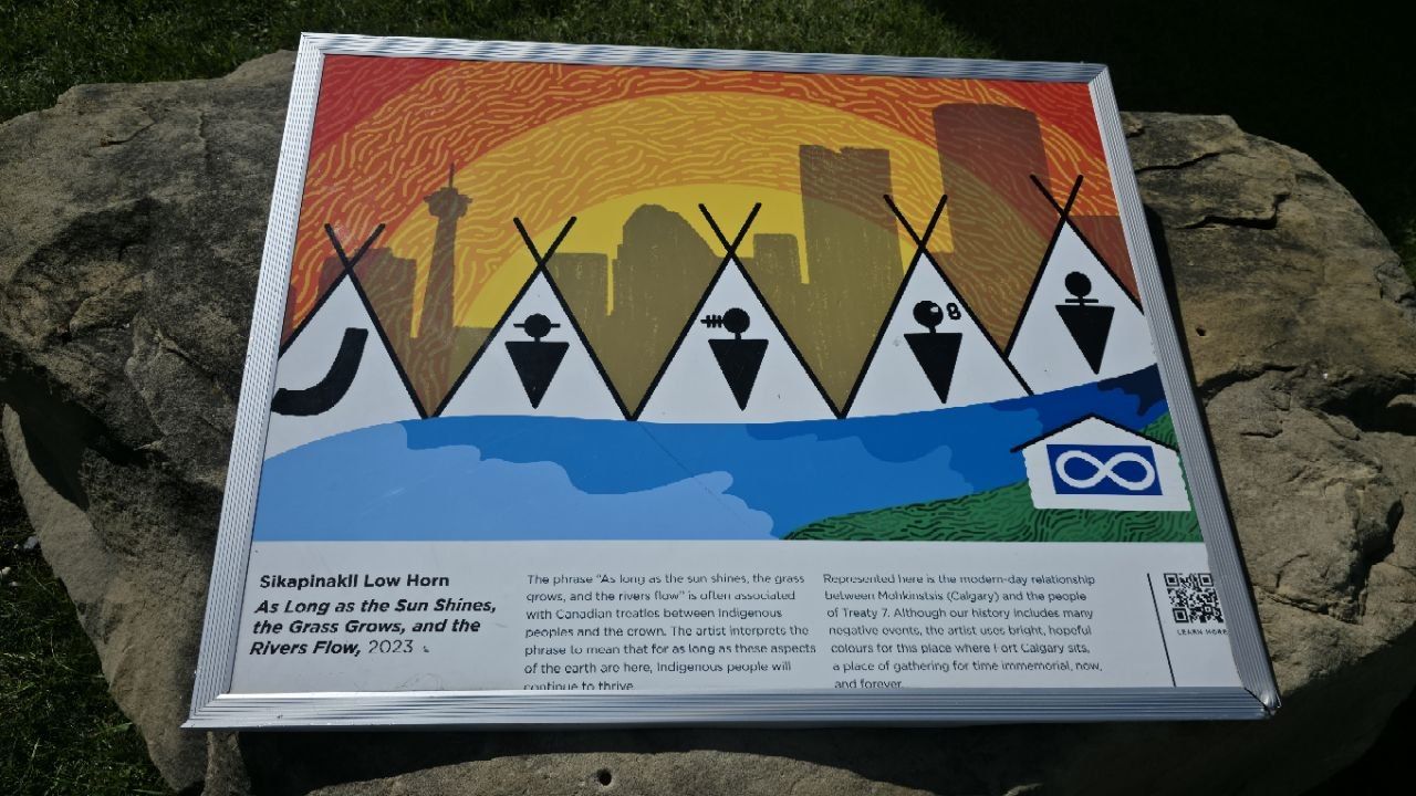 A framed panel on a boulder: five white tipis with black symbols in front of a sunburst Calgary skyline, a blue river below, a Métis infinity flag; titled 'As Long as the Sun Shines, the Grass Grows, and the Rivers Flow', Sikapinakii Low Horn, 2023.