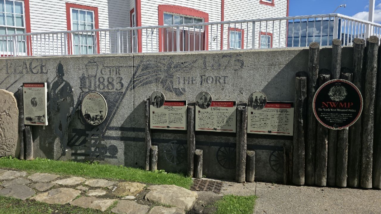 A long concrete timeline wall with sandblasted lettering 'PEACE', 'CPR 1883', '1875 THE FORT', a cut-steel silhouette of a Mountie, wagon wheels, and a row of interpretive plaques; a white clapboard house with red trim above.