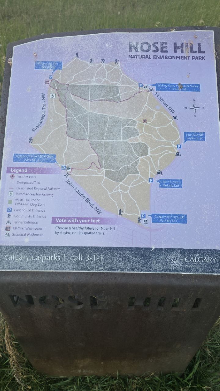 A rusted steel kiosk with a faded park map: Nose Hill Natural Environment Park, trails, parking lots, off-leash zones, washrooms; a legend with no symbol for anything archaeological; 'Vote with your feet'.