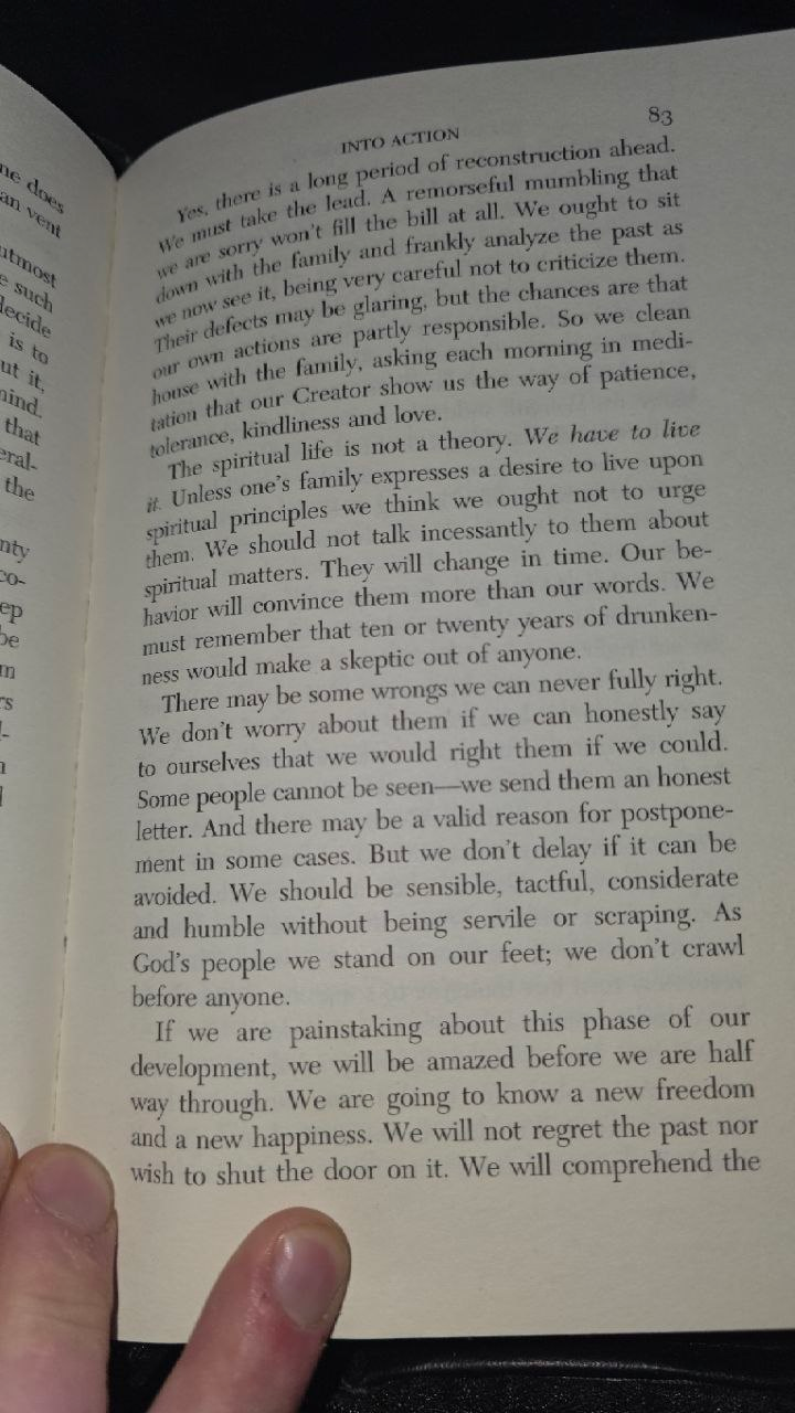 Page 83 of the Big Book photographed at night, a thumb holding the page flat; the Promises begin in the last paragraph: 'If we are painstaking about this phase of our development...'