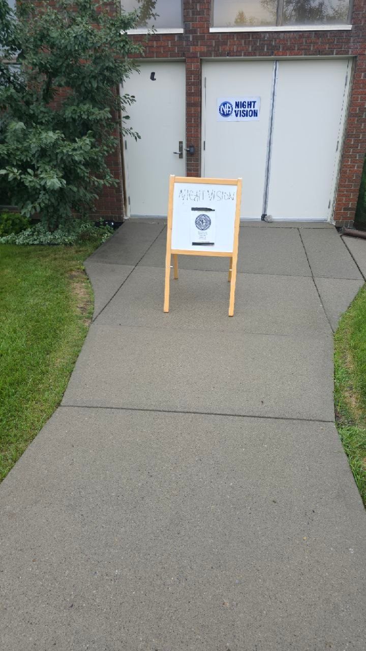A concrete walk leading to two white steel doors in a brick wall; a blue NA 'Night Vision' sticker at eye height on the right-hand door, a rubber wedge on the ground beside it; in the middle of the walk a wooden easel holds a whiteboard hand-lettered NIGHT VISION in marker with the NA logo taped beneath; an evening overcast sky, a shrub, mown grass.