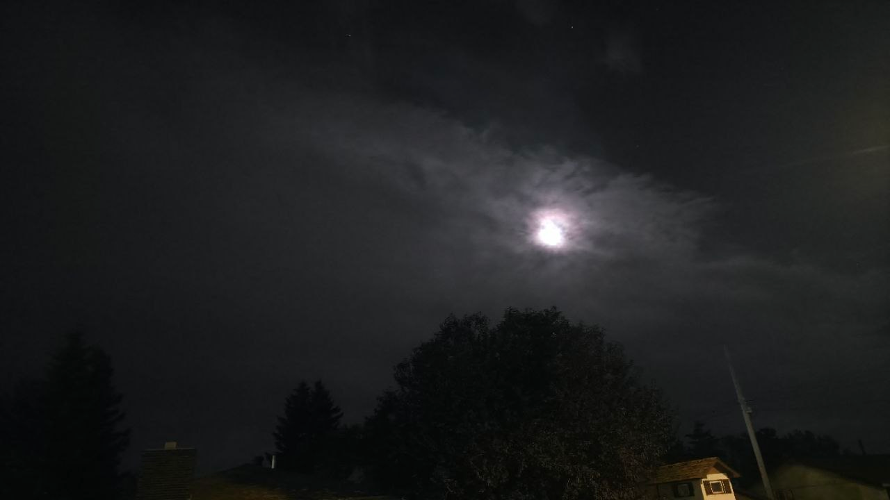 The moon breaking through a heavy cloud deck, a soft corona bleeding into the vapor, a large tree and rooflines silhouetted below.