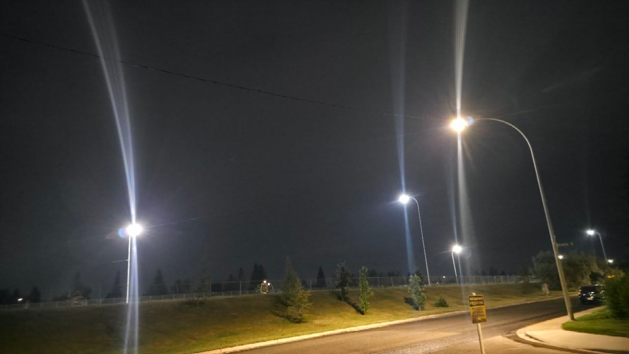 A dark suburban street in Calgary at night: four streetlights on gooseneck poles throw tall lens-flare arcs upward, a chain-link fence runs along a grassy berm with young trees, a single power line crosses the top of the frame.