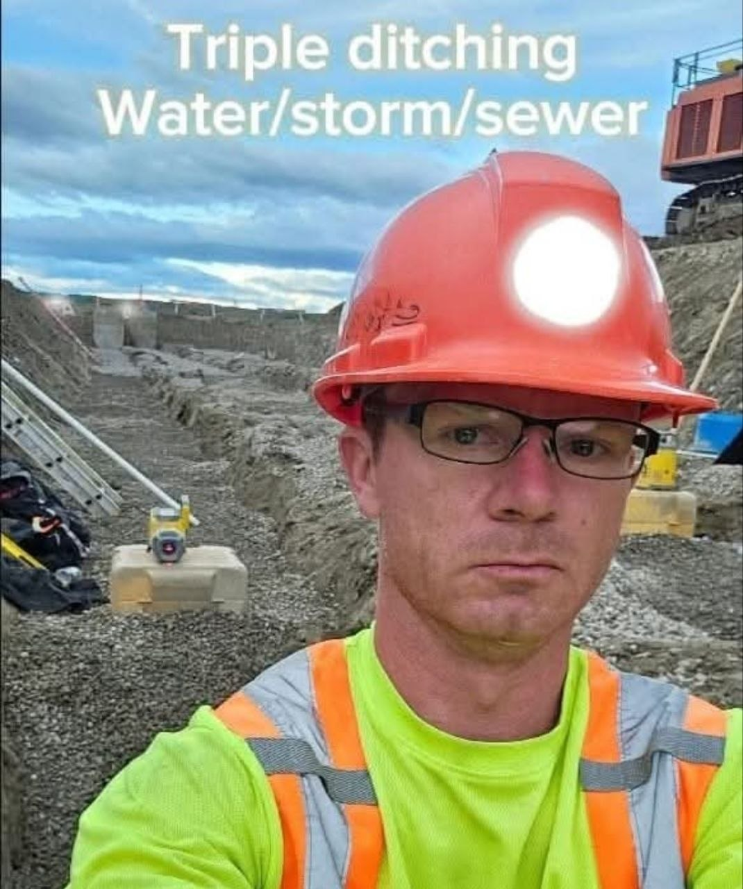 Selfie of Derick in an orange hard hat and hi-vis shirt standing in a deep excavation; behind him a pipe laser on its case sights down a gravel-bedded trench toward a distant manhole; ladders lean against the cut wall; an excavator sits on the bench above against a blue prairie sky.